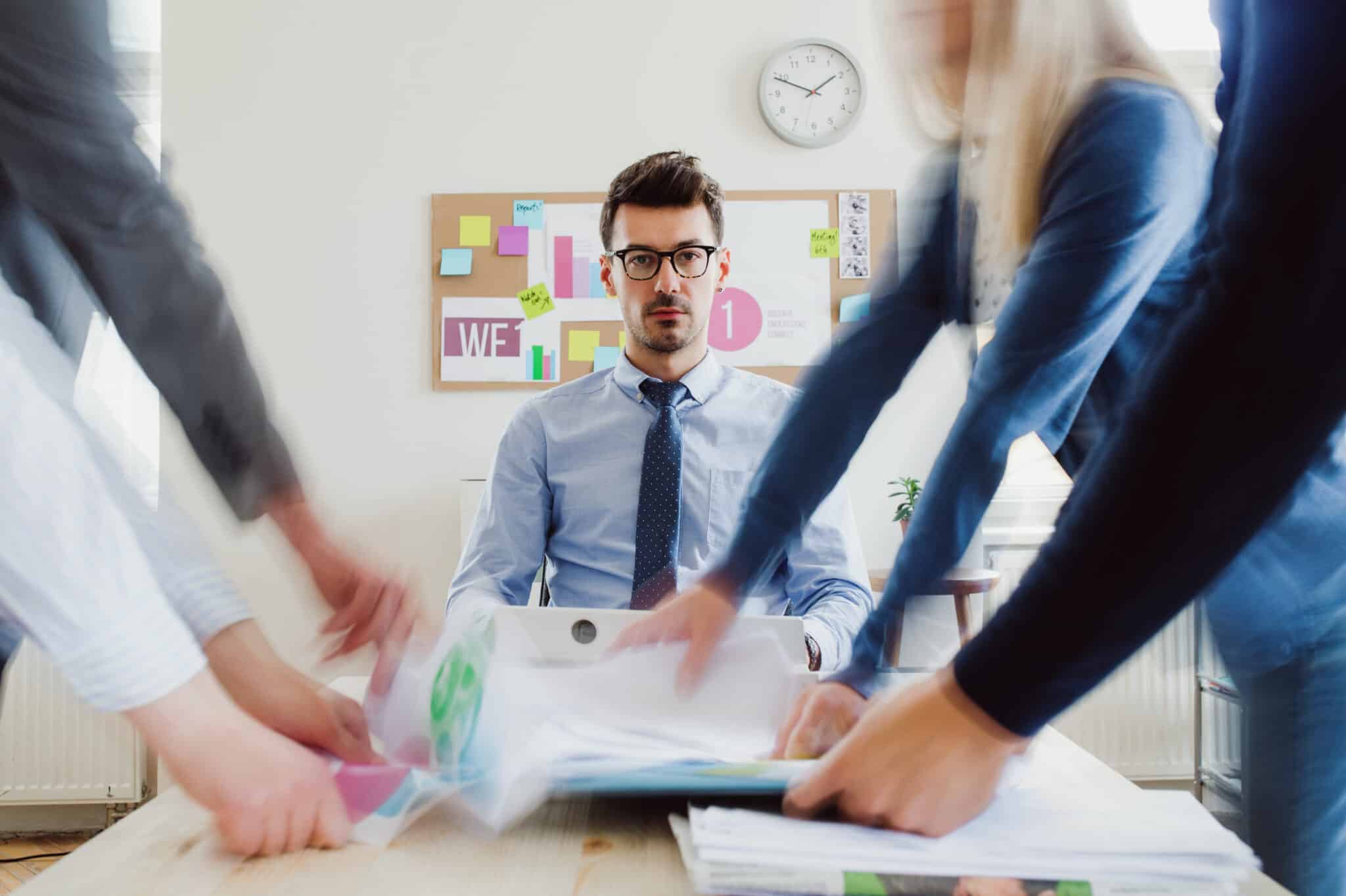 A man sits at a desk looking towards the camera while blurred figures move around him carrying paperwork in a busy office.