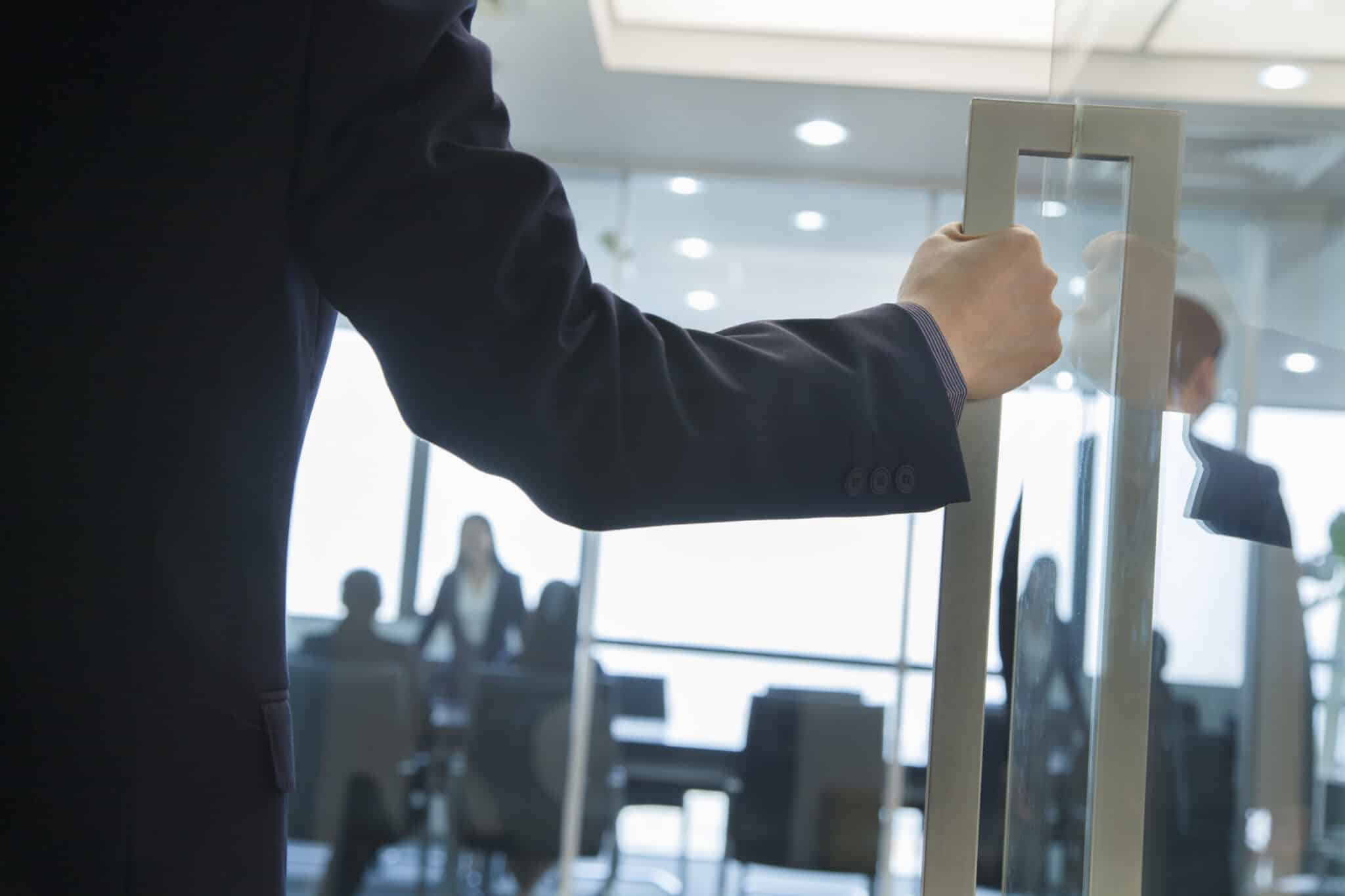 Person in a dark suit opening a glass door into a bright office meeting room, with several people visible in the background.