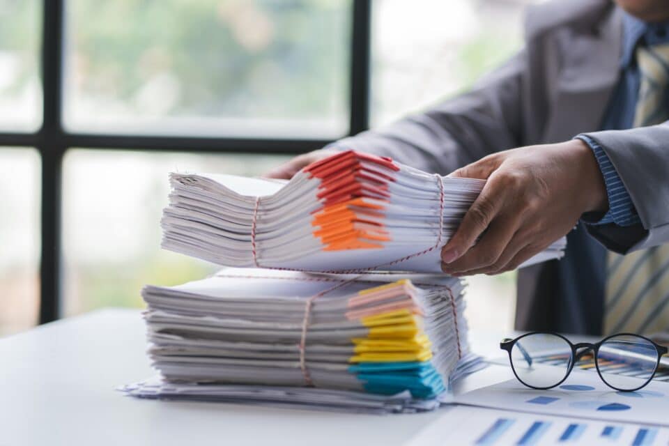 A person in a suit places a thick bundle of paperwork onto a large stack of documents with colourful tabs on an office desk.